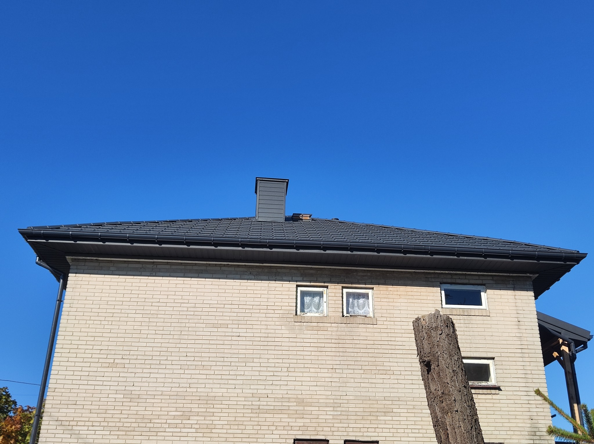 Dom z jasnej cegły z ciemnym dachem i rynnami na tle błękitnego nieba, widoczne okna z koronkowymi firankami.