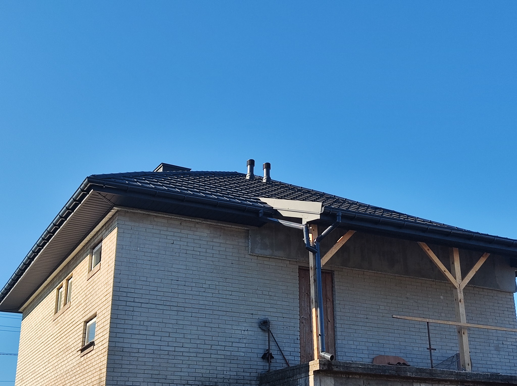 Dom z szarej cegły z nowym, ciemnym dachem z rynnami, widoczne kominy i fragment rusztowania.