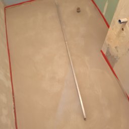 TD-Remonty - Wylewka samopoziomująca w łazience przed położeniem płytek, widoczny odpływ liniowy i listwa poziomująca oraz fragment stopy w obuwiu roboczym.