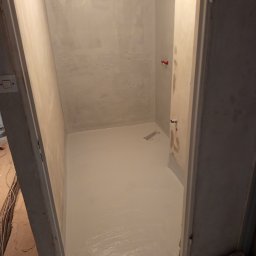 TD-Remonty - Świeżo pomalowane, jasnoszare pomieszczenie z widocznymi śladami pędzla na podłodze i ścianach, surowy stan wykończenia, widoczne instalacje hydrauliczne