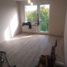 TD-Remonty - Pokój po remoncie z nową podłogą z paneli, widoczne narzędzia i odkurzacz, światło słoneczne wpadające przez okno z widokiem na zieleń.