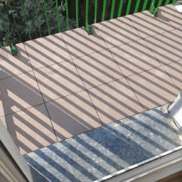 TD-Remonty - Brązowe płytki ułożone na balkonie, cień od zielonej balustrady tworzy geometryczny wzór, widoczny fragment progu i drewniana podłoga wewnątrz pomieszczenia.