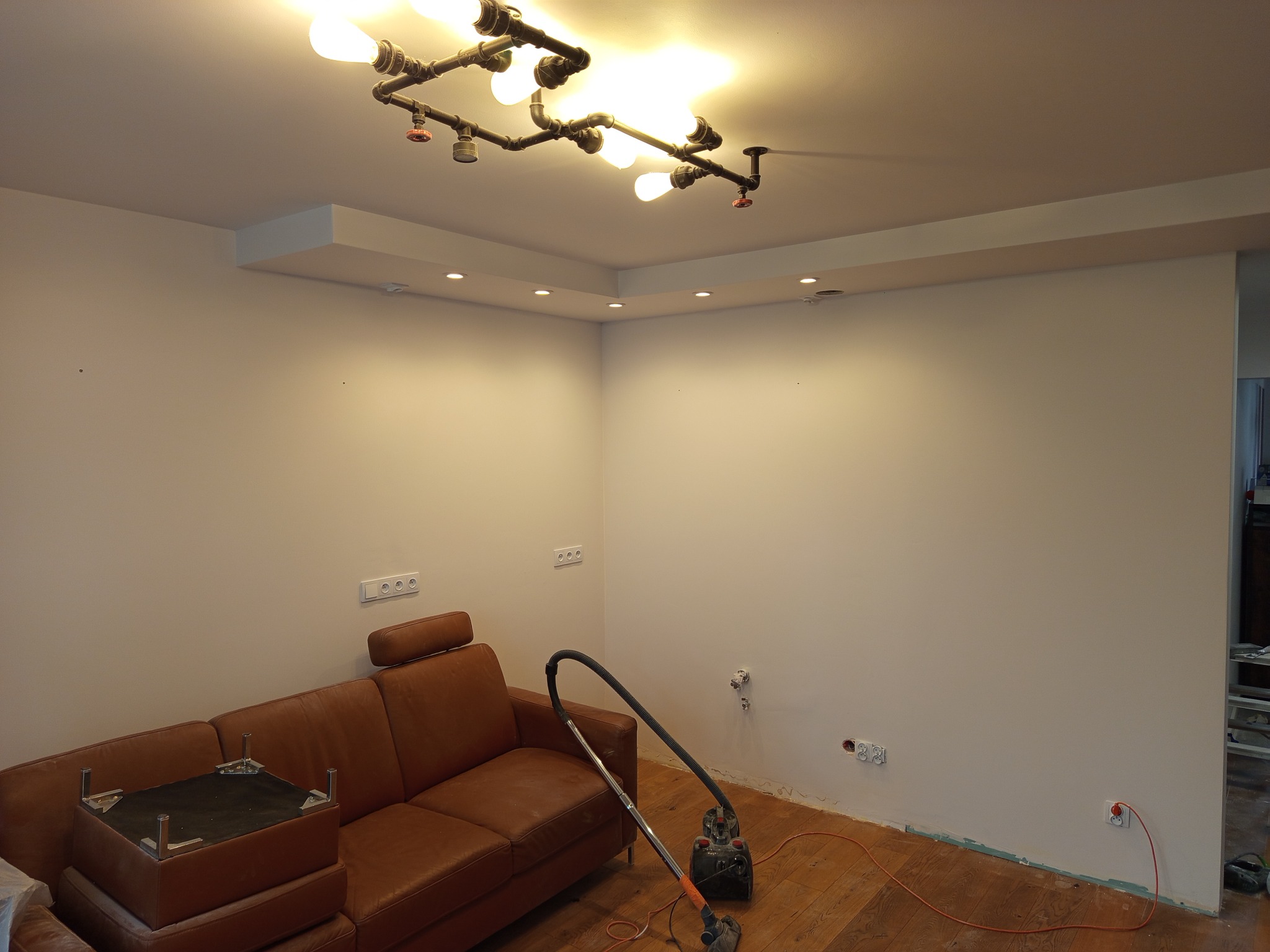 Salon w trakcie porządkowania po pracach wykończeniowych, widoczna skórzana sofa, odkurzacz i nietypowa lampa sufitowa z rur w stylu industrialnym.
