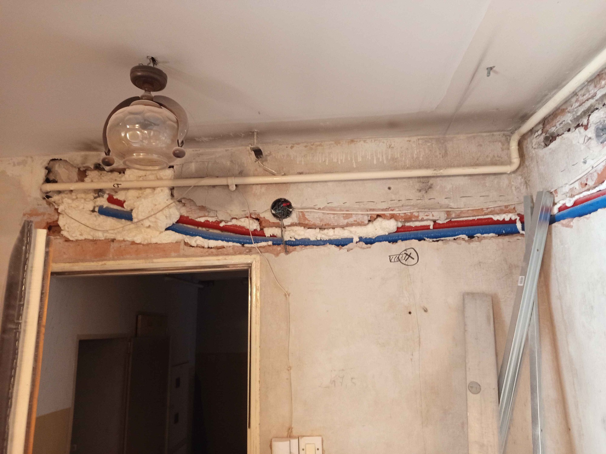 Ściana w trakcie remontu z odsłoniętymi rurami grzewczymi zabezpieczonymi pianką, widoczne ślady po starych instalacjach, wisząca lampa w stylu retro i częściowo usunięty tynk.