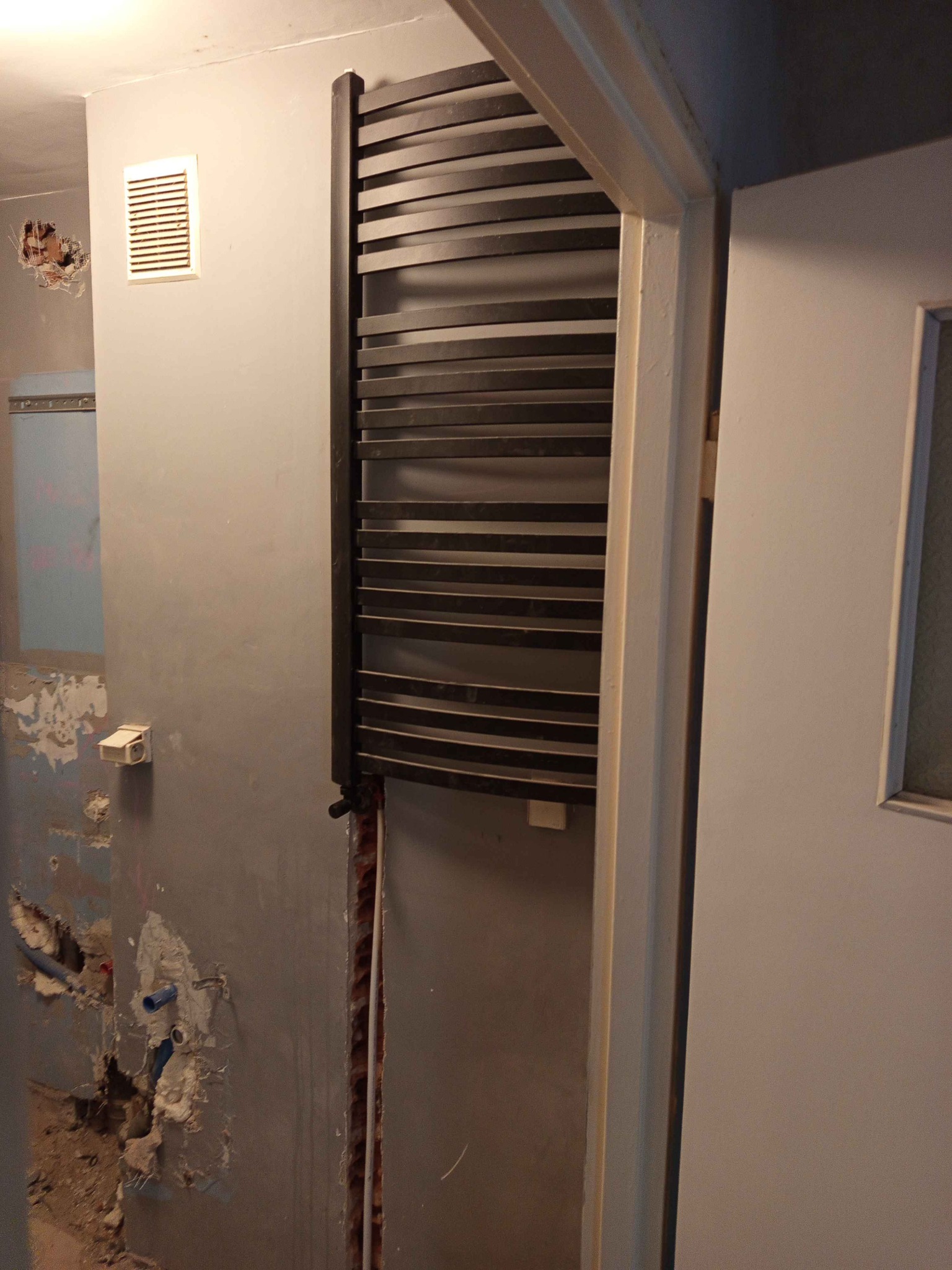 Montaż czarnego grzejnika drabinkowego w trakcie remontu łazienki, widoczne ślady po kuciu w ścianie i niezabezpieczone rury.