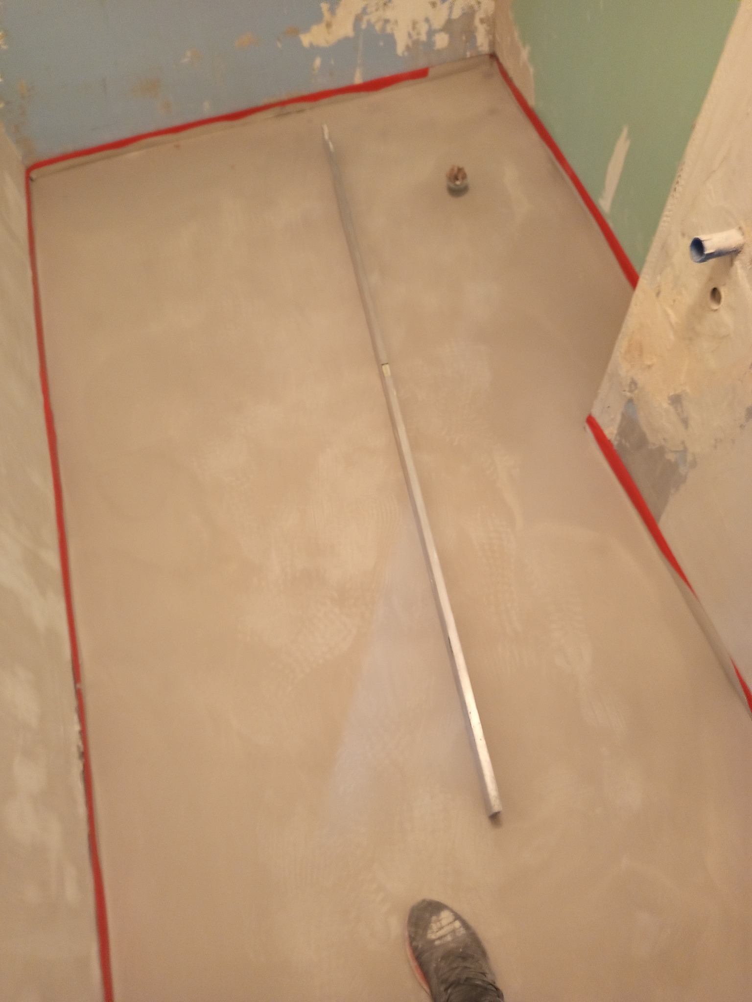 Wylewka samopoziomująca w łazience przed położeniem płytek, widoczny odpływ liniowy i listwa poziomująca oraz fragment stopy w obuwiu roboczym.