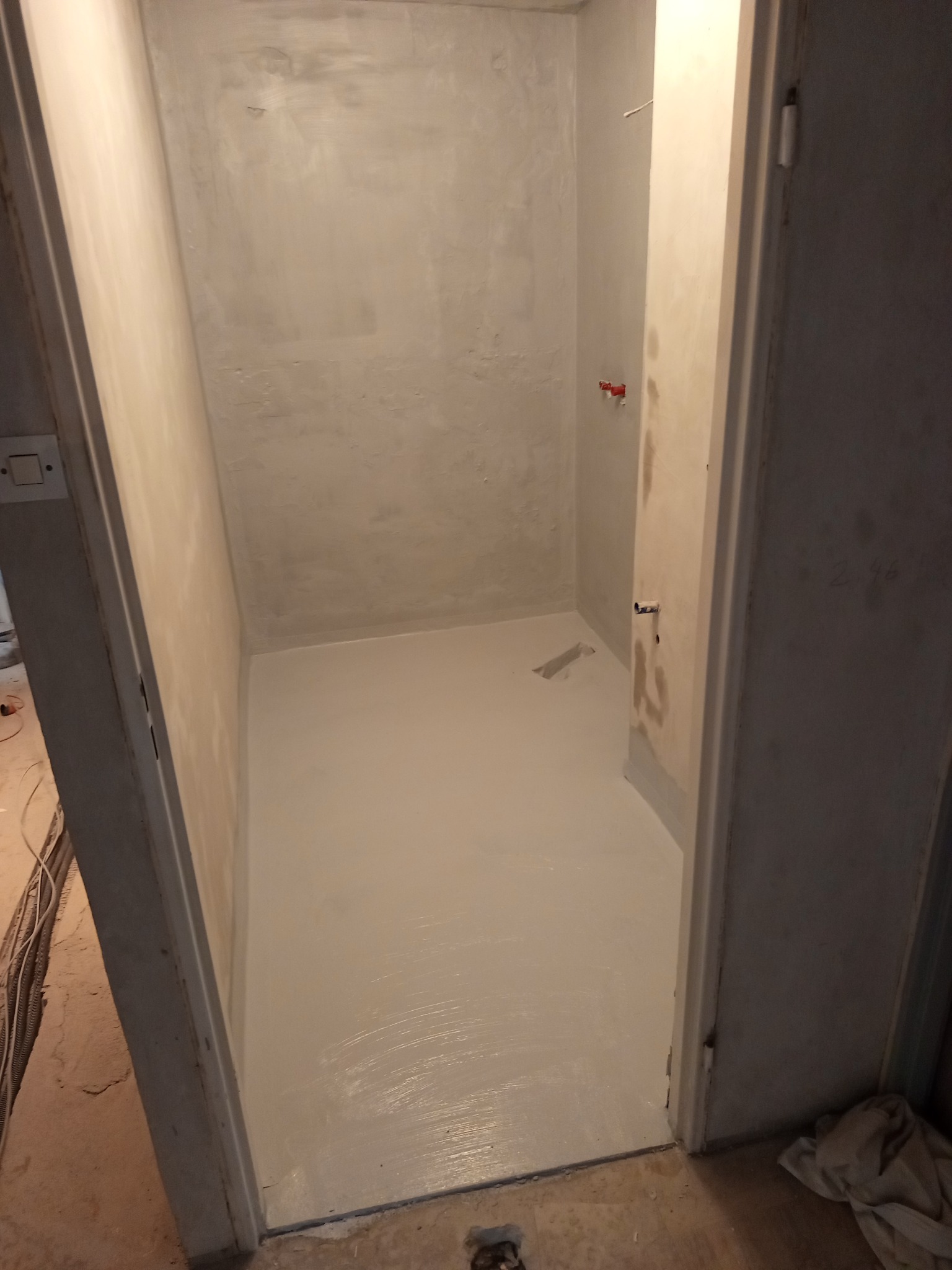 Świeżo pomalowane, jasnoszare pomieszczenie z widocznymi śladami pędzla na podłodze i ścianach, surowy stan wykończenia, widoczne instalacje hydrauliczne