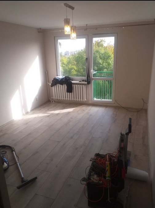 Pokój po remoncie z nową podłogą z paneli, widoczne narzędzia i odkurzacz, światło słoneczne wpadające przez okno z widokiem na zieleń.