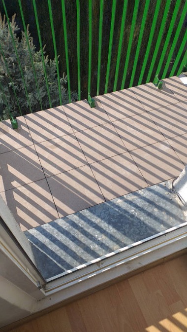 Brązowe płytki ułożone na balkonie, cień od zielonej balustrady tworzy geometryczny wzór, widoczny fragment progu i drewniana podłoga wewnątrz pomieszczenia.