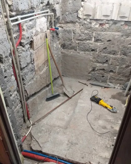 Przygotowanie podłoża w łazience do dalszych prac: surowe ściany z kamienia, wystające rury, szczotka, szpadel i szlifierka kątowa na podłodze.