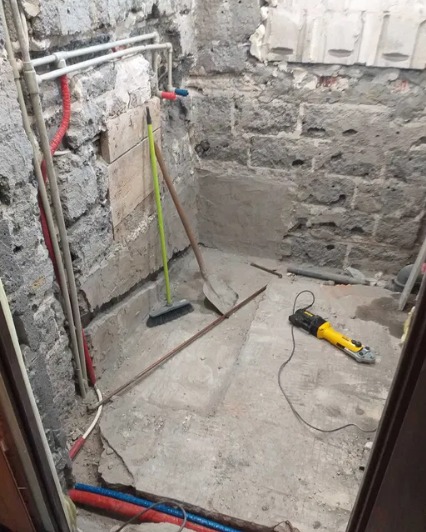 Surowa łazienka w trakcie remontu: odsłonięte cegły, nowe rury, szpachelka i szczotka oparta o ścianę, szlifierka kątowa na podłodze.