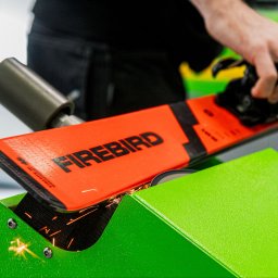 Magdalena Ramach-Nowak - Ostrzenie pomarańczowej narty Firebird na zielonej maszynie, z iskrami i dłonią operatora w tle.