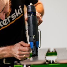 Magdalena Ramach-Nowak - Mężczyzna w czarnej koszulce z logo 'bikerski' naprawia ślizg narty za pomocą specjalistycznego narzędzia z niebieskimi elementami.