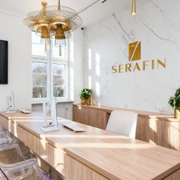 Magdalena Ramach-Nowak - Eleganckie wnętrze biura z recepcją: drewniane biurko, przezroczyste krzesła, marmurowa ściana z logo firmy 'Serafin', złota lampa wisząca i rośliny doniczkowe.