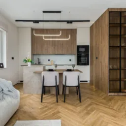 Nowoczesna kuchnia z wyspą i drewnianą zabudową, oświetlona lampami LED. Parkiet w jodełkę, szara sofa z kocem w tle. Minimalistyczny design.