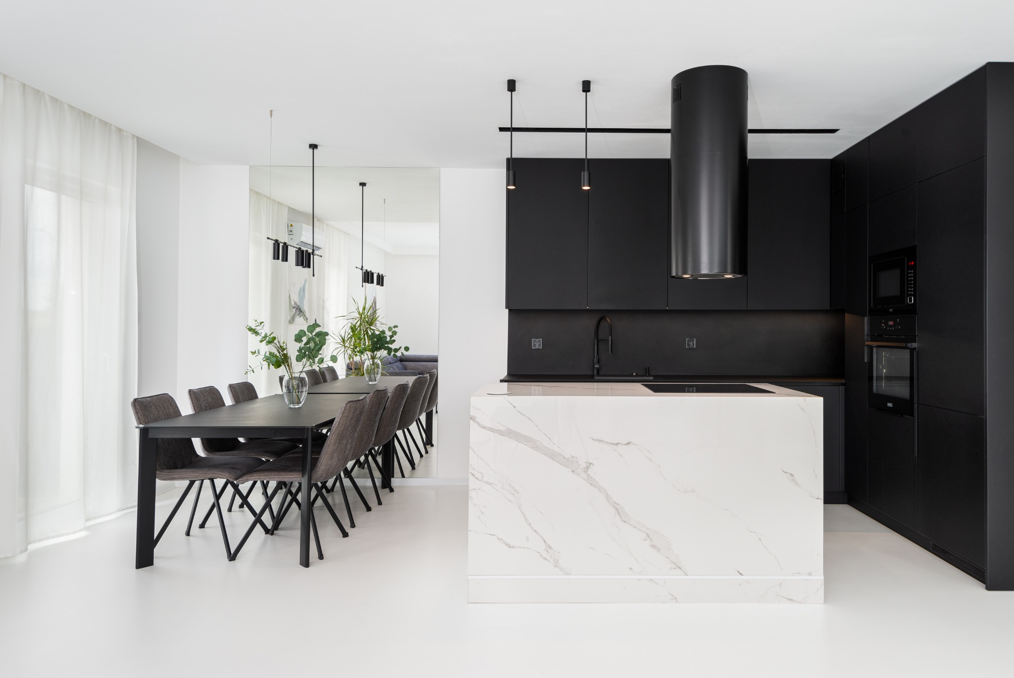 Nowoczesna kuchnia z czarnymi szafkami, białą wyspą z marmurowym wzorem i długim stołem jadalnym z szarymi krzesłami, odbijającym się w lustrze.