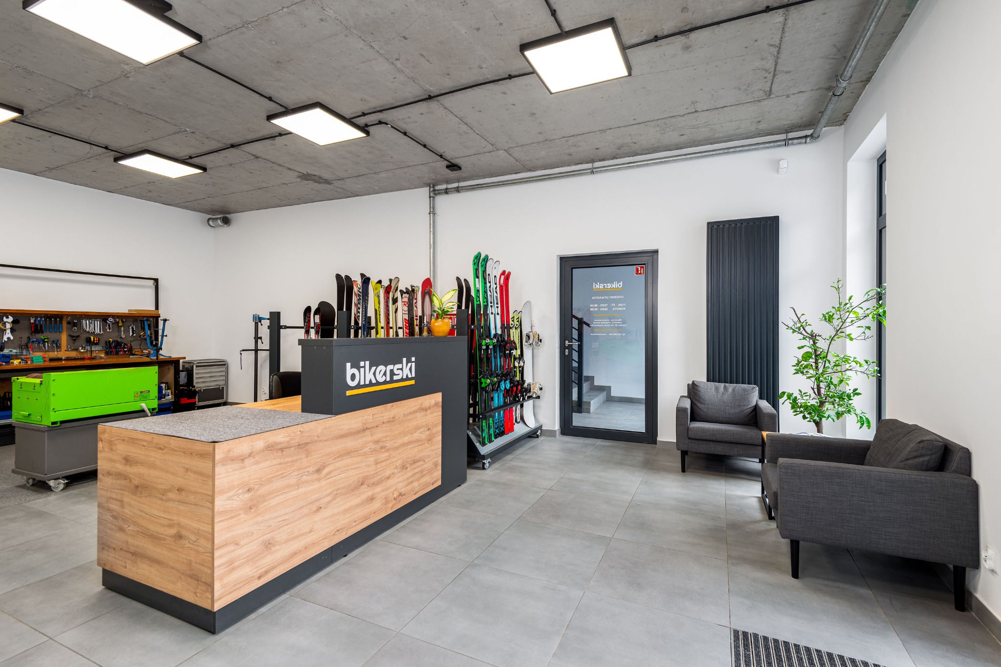 Wnętrze serwisu narciarskiego: recepcja z logo 'bikerski', ekspozycja nart, stanowisko serwisowe z narzędziami, szare fotele dla klientów, surowy betonowy sufit z lampami.