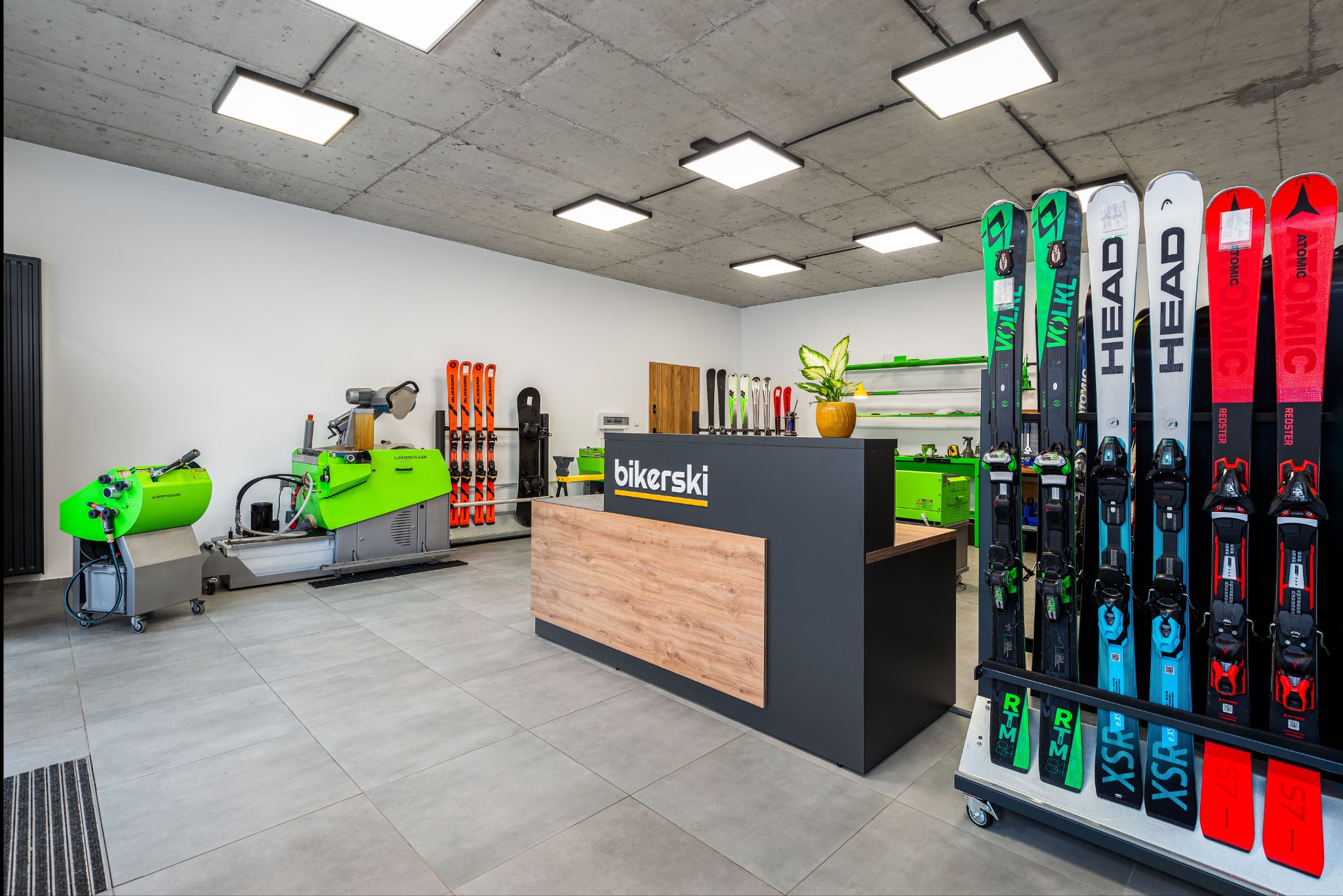 Wnętrze sklepu sportowego Bikerski z maszynami do serwisowania nart Wintersteiger, ekspozycją nart marek Volkl, Head i Atomic oraz ladą recepcyjną z logo.