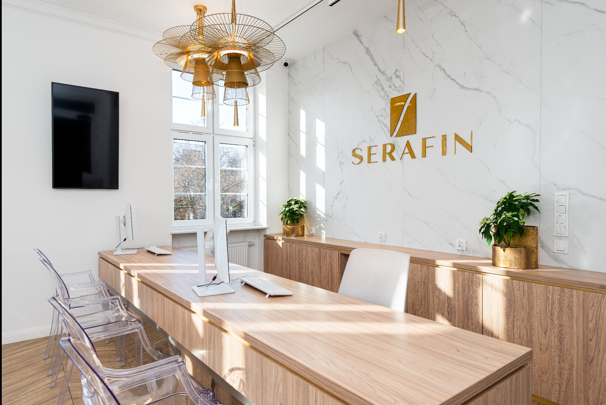 Eleganckie wnętrze biura z recepcją: drewniane biurko, przezroczyste krzesła, marmurowa ściana z logo firmy 'Serafin', złota lampa wisząca i rośliny doniczkowe.