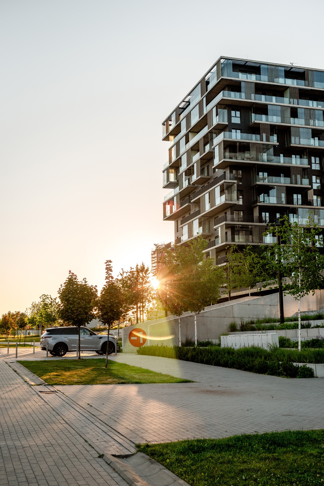 Nowoczesny budynek apartamentowy z balkonami, widok z poziomu ulicy, zieleń miejska i zaparkowany srebrny SUV w promieniach zachodzącego słońca.