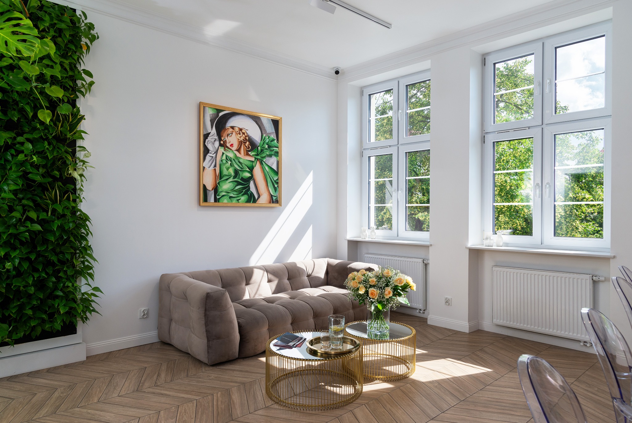 Jasne wnętrze z beżową sofą, dwoma okrągłymi stolikami kawowymi ze złotymi podstawami, pionową ścianą z roślin, obrazem kobiety w zielonej sukni i parkietem ułożonym w jodełkę.