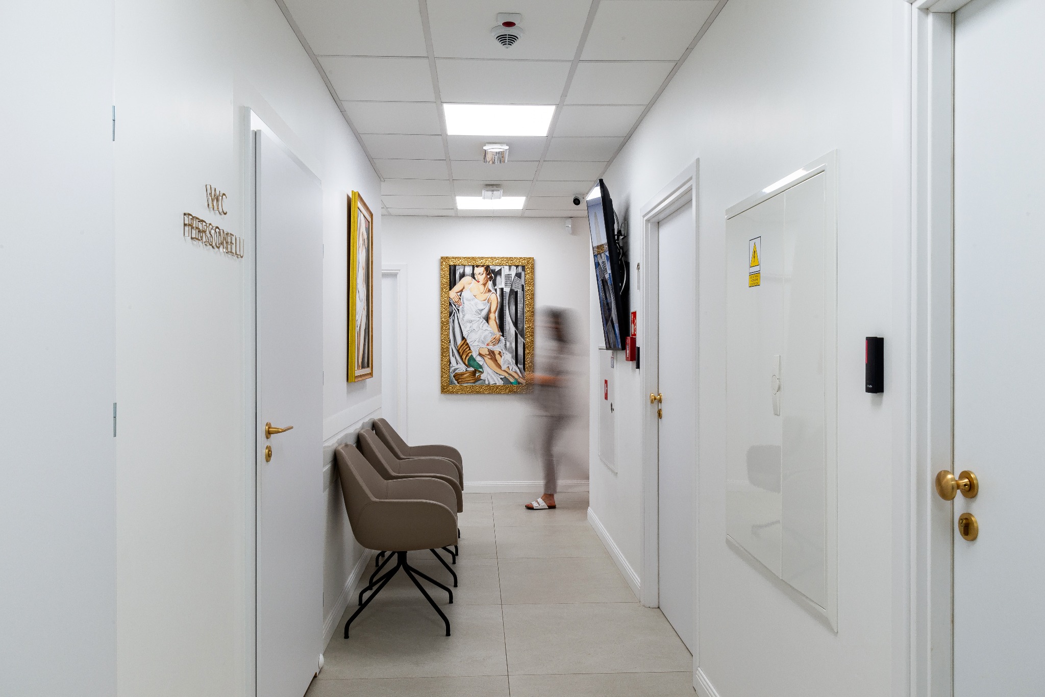 Korytarz w jasnych barwach z trzema krzesłami na czarnych nogach, obrazami w złotych ramach, telewizorem, przechodzącą osobą i tabliczką informacyjną na drzwiach.