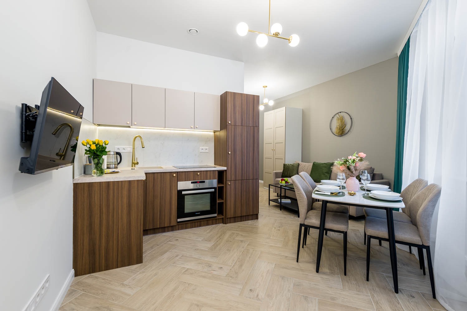 Nowoczesne wnętrze mieszkania z aneksem kuchennym: drewniane szafki, złota armatura, stół z nakryciem i bukietem kwiatów, telewizor na ścianie.