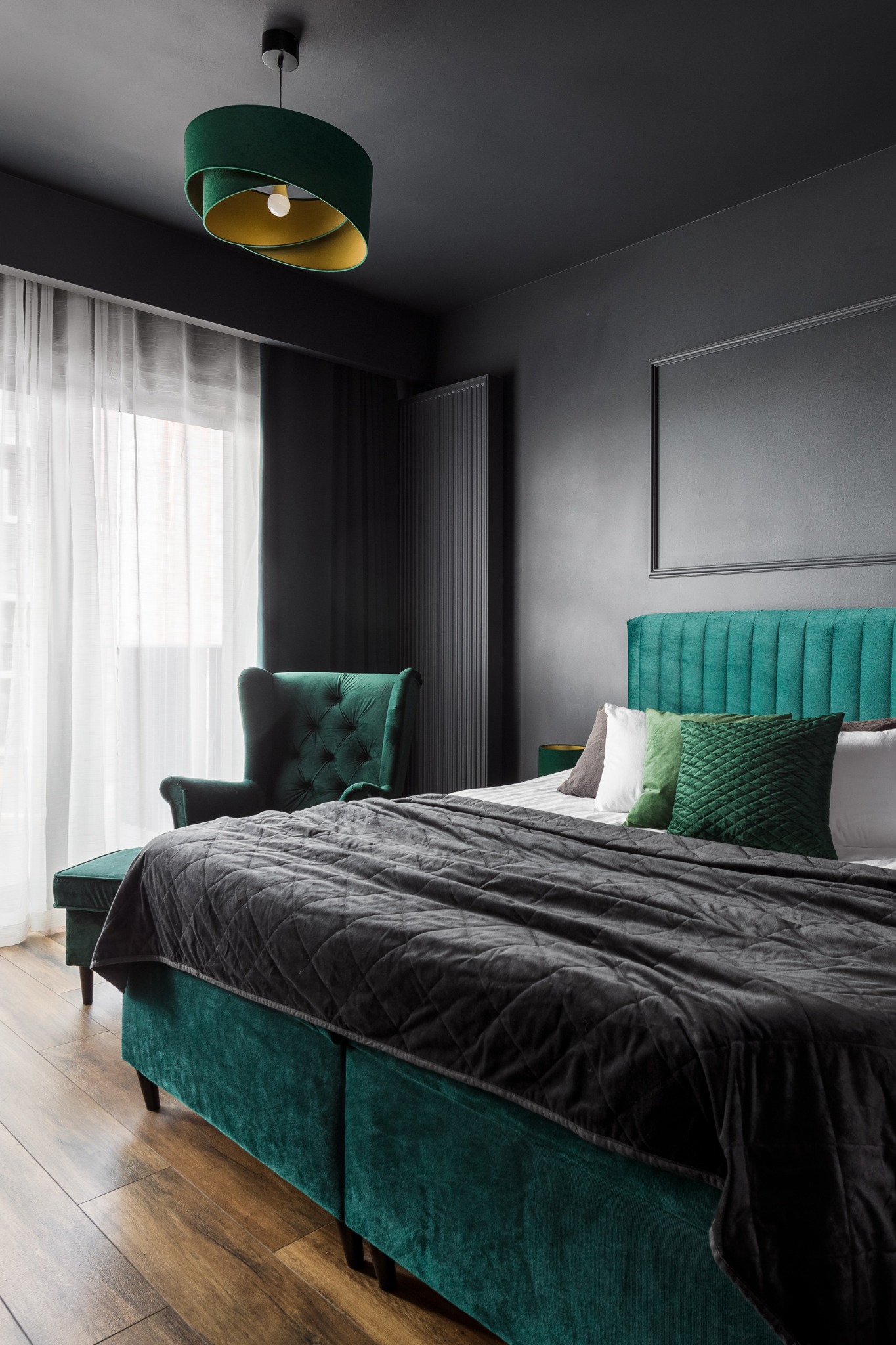 Ciemnoszara sypialnia z akcentami butelkowej zieleni: tapicerowane łóżko z pikowanym pledem, fotel typu uszak i okrągła lampa z elementami złota.