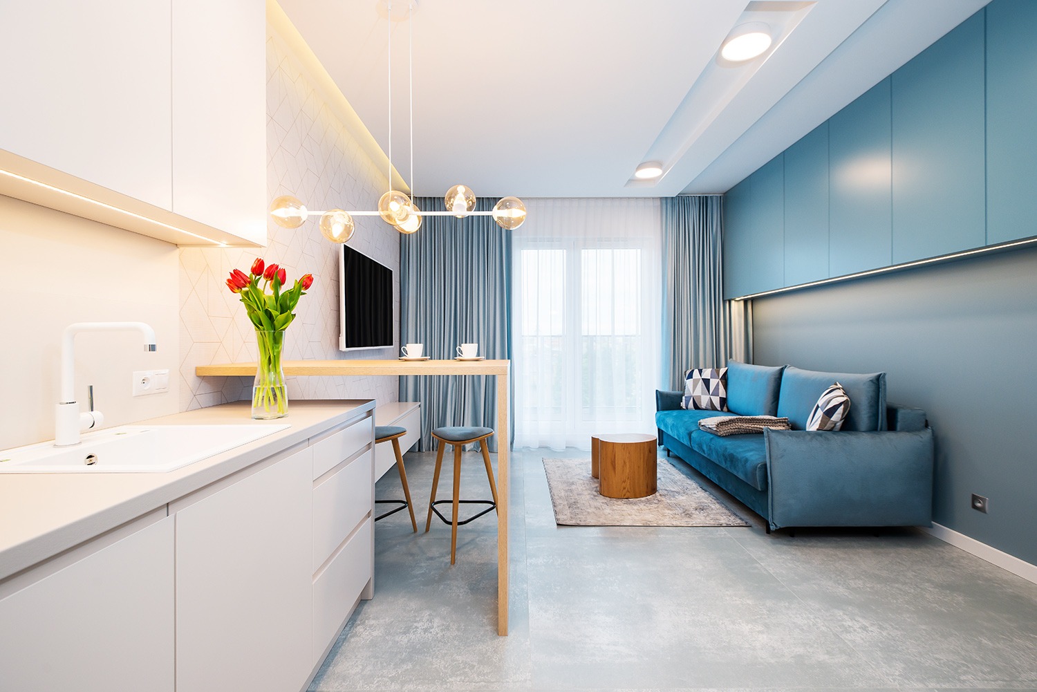 Jasne wnętrze mieszkania z otwartą kuchnią, błękitną ścianą i turkusową sofą, tulipany w wazonie na blacie.