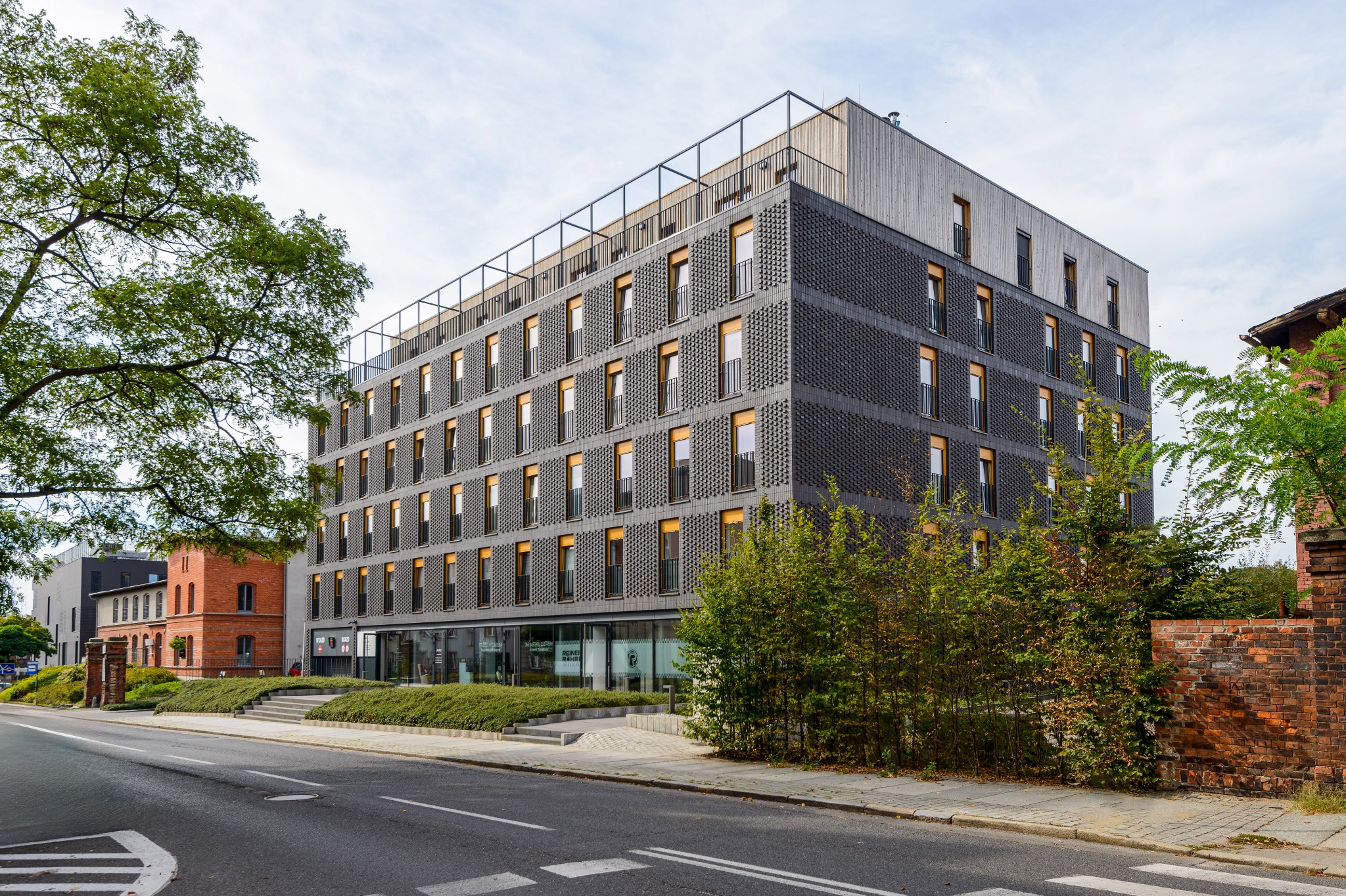 Nowoczesny budynek biurowy z ciemną elewacją o fakturze cegły, z drewnianą nadbudówką i metalową balustradą na dachu, widziany z poziomu ulicy.