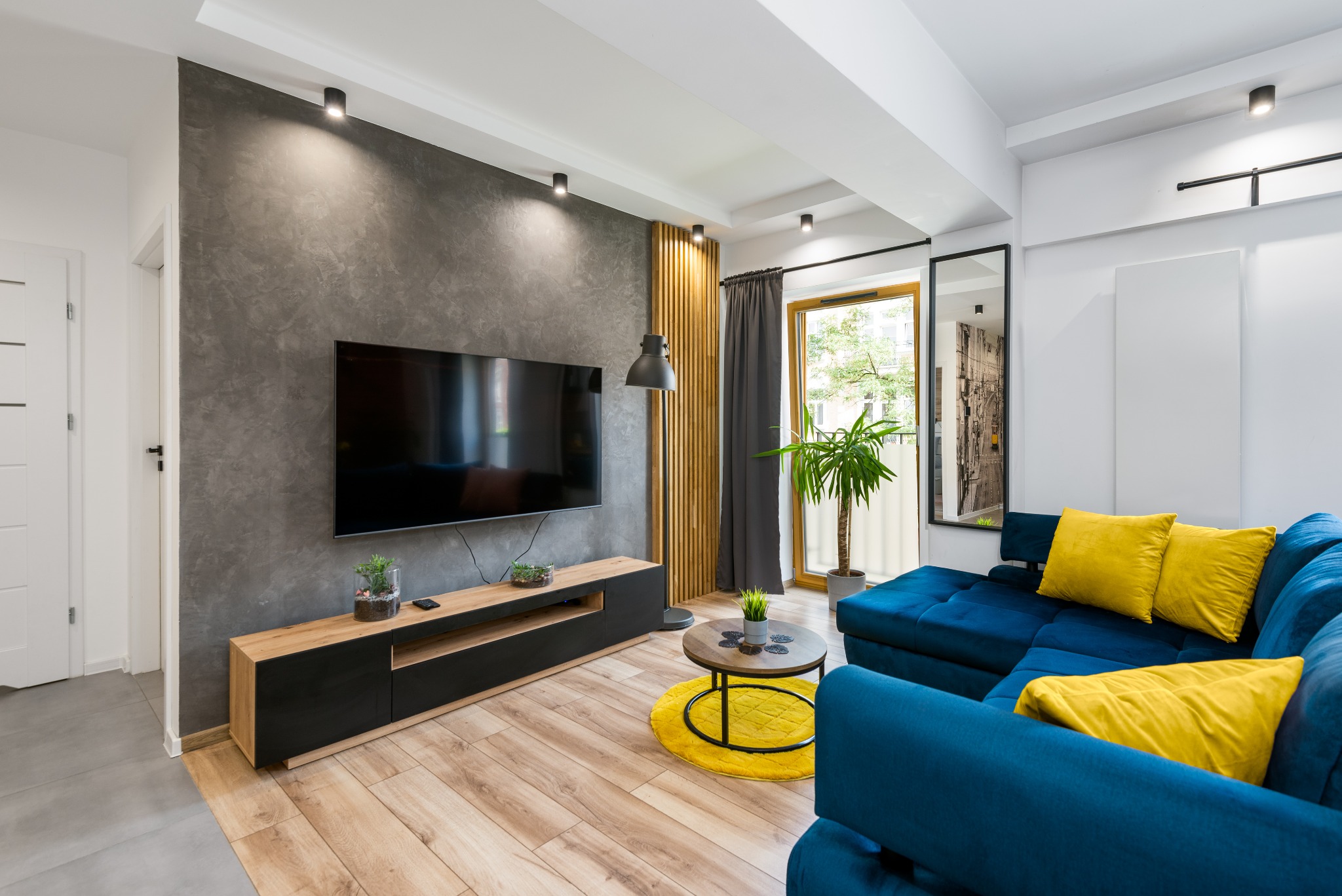 Nowoczesny salon z szarą ścianą z efektem betonu, drewnianą podłogą, niebieską sofą z żółtymi poduszkami, telewizorem i roślinami w doniczkach. Widoczne elementy dekoracyjne: okrągły stolik...