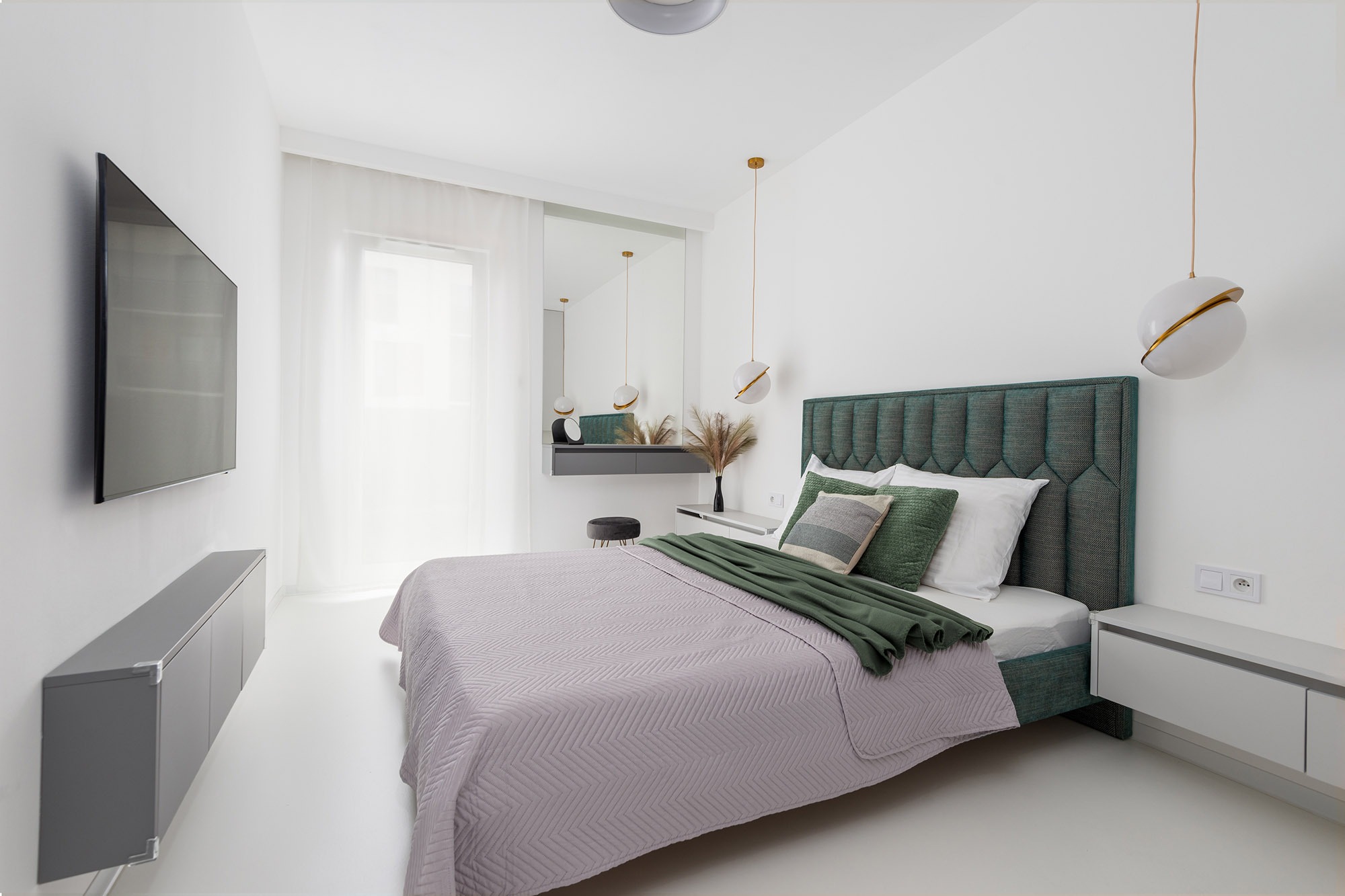Nowoczesna sypialnia z łóżkiem z zielonym zagłówkiem, telewizorem na ścianie i wiszącą toaletką z lustrem, oświetlona kulistymi lampami.
