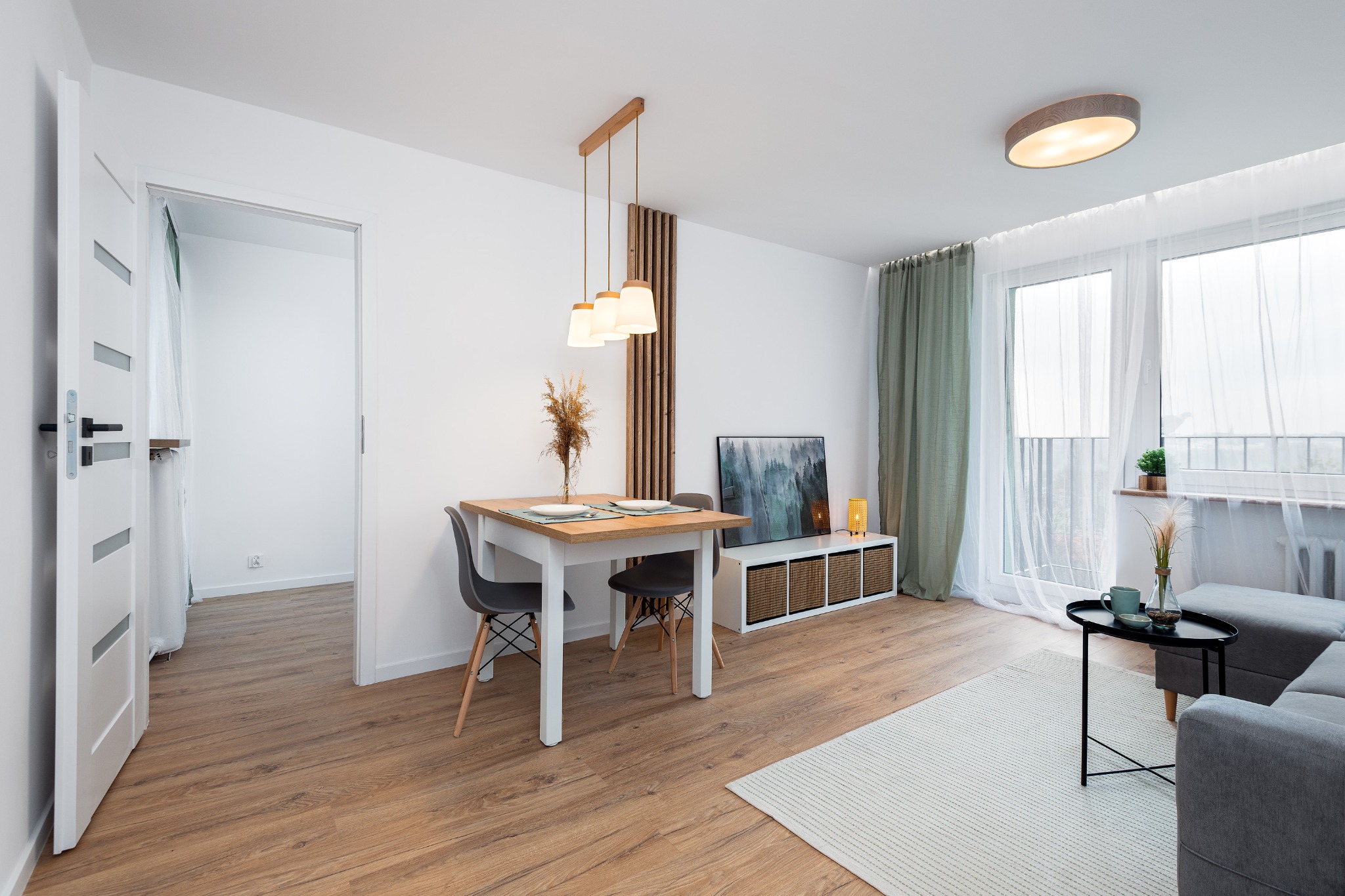 Jasne wnętrze mieszkania z drewnianą podłogą, białymi ścianami, stołem z nakryciem, szarą sofą i dużym oknem z firanami.