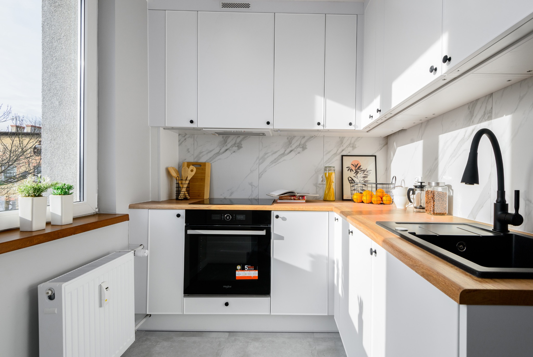 Jasna kuchnia z białymi szafkami, drewnianym blatem i czarną armaturą, oświetlona naturalnym światłem wpadającym przez okno, z widocznym grzejnikiem pod oknem.
