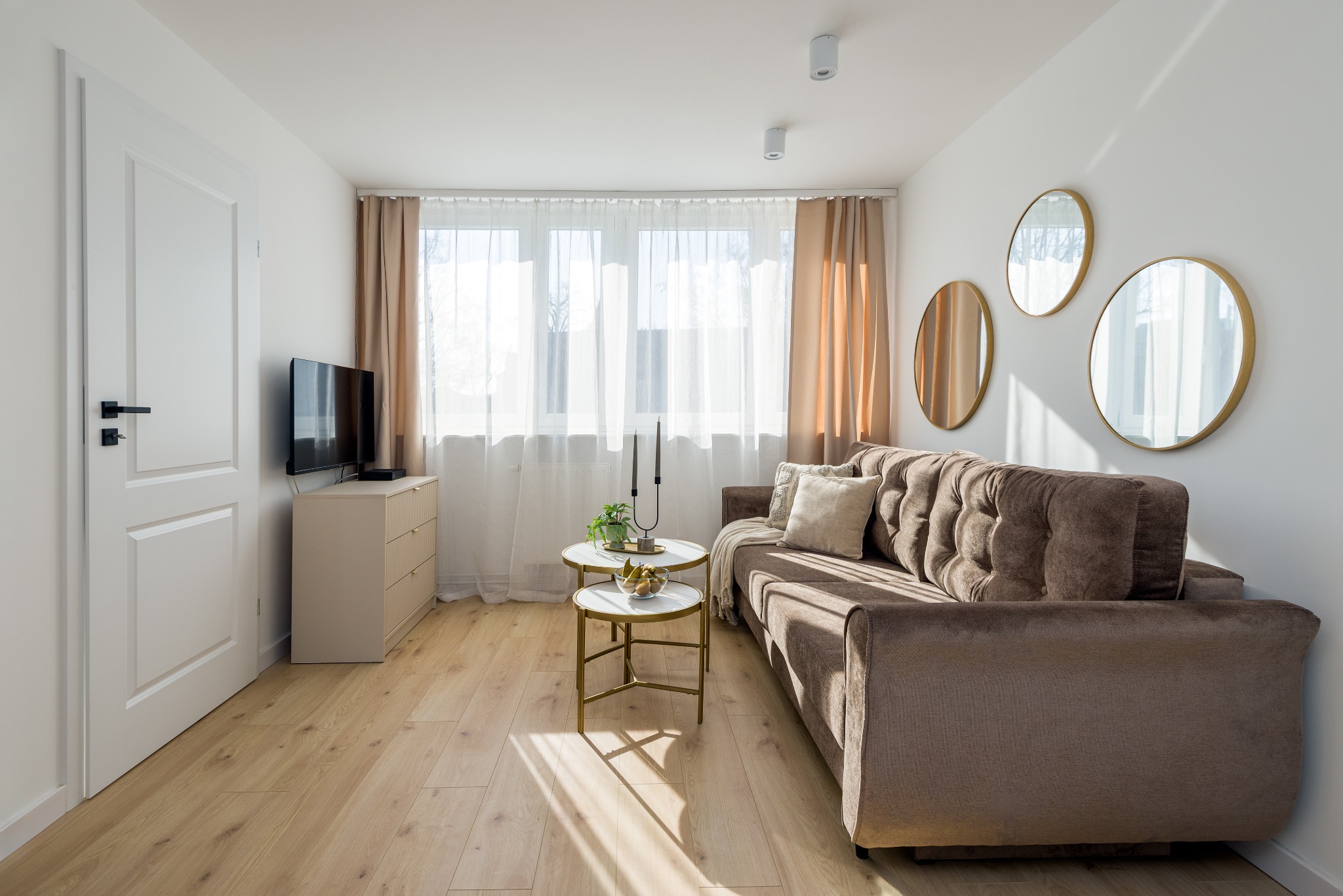 Jasny salon z brązową sofą, okrągłymi lustrami na ścianie, stolikami kawowymi i drewnianą podłogą, oświetlony naturalnym światłem wpadającym przez okno z białymi zasłonami.