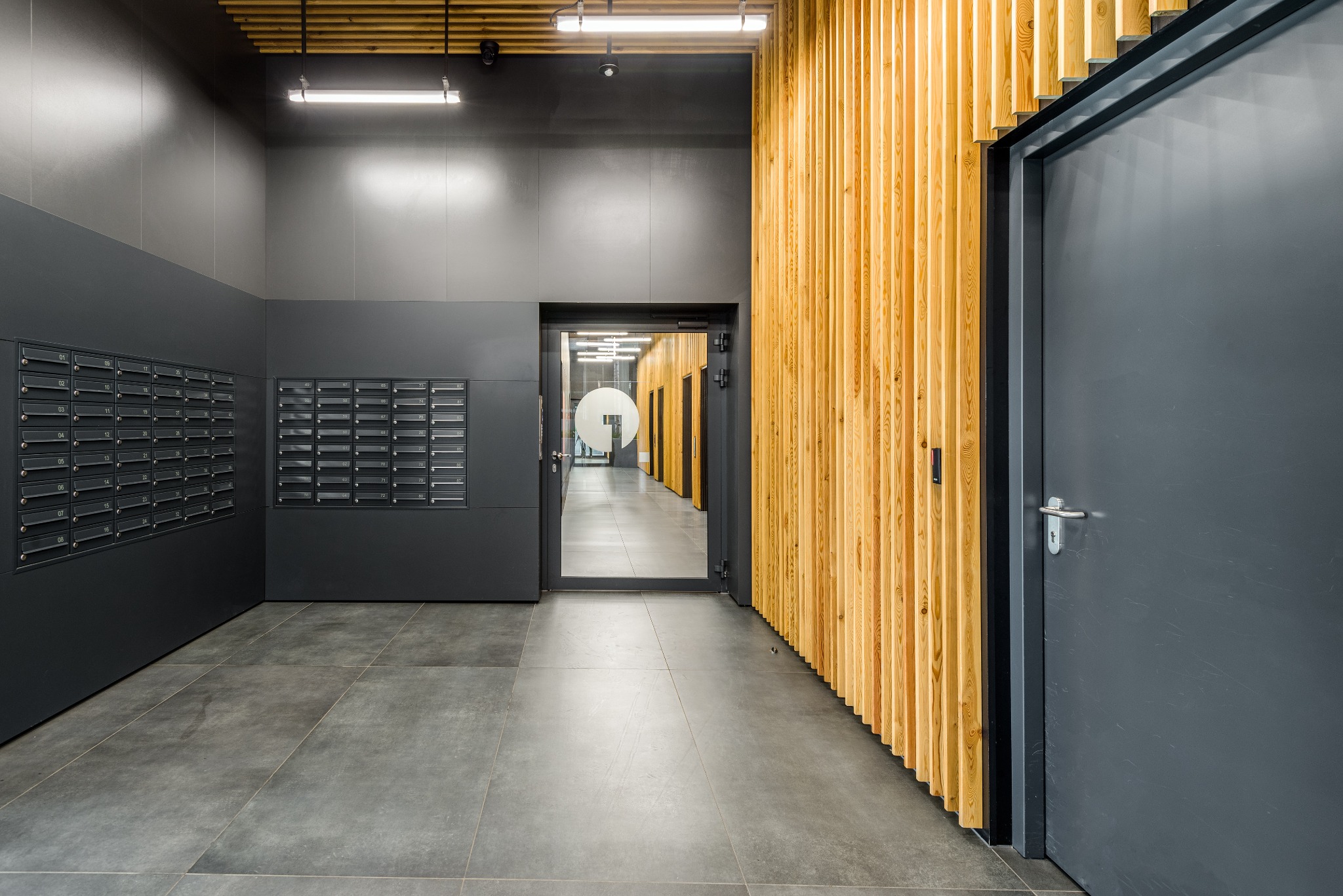 Wnętrze nowoczesnego holu wejściowego z szarymi ścianami, drewnianą okładziną, rzędami skrzynek pocztowych i widokiem na korytarz z przeszklonymi drzwiami.