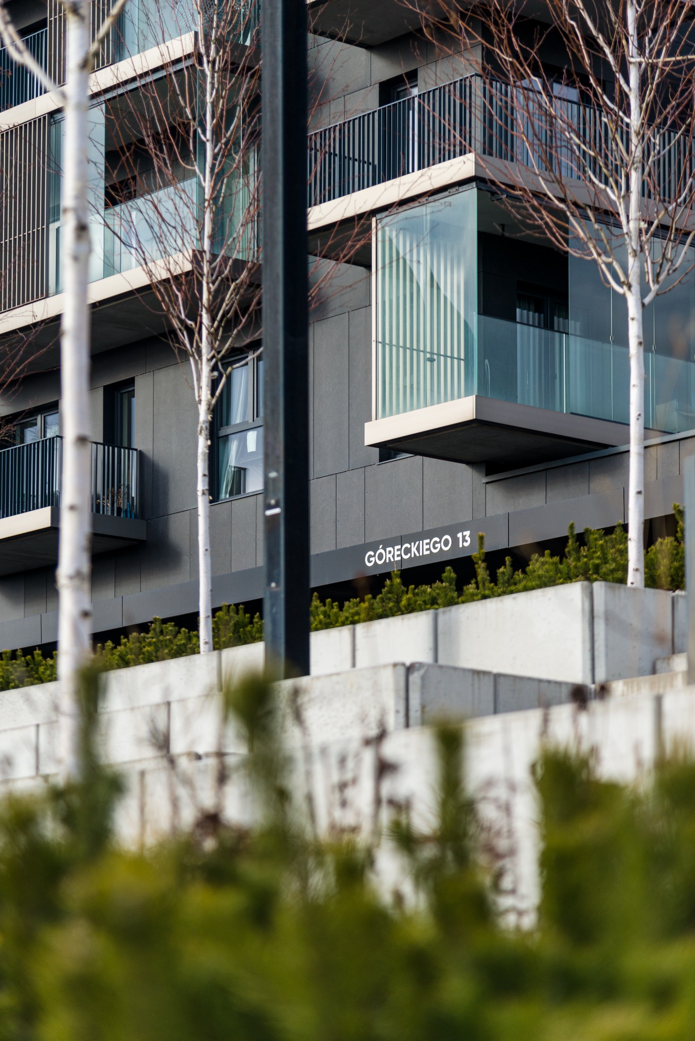 Nowoczesny budynek mieszkalny z ciemną elewacją i szklanymi balkonami, widoczny adres 'Góreckiego 13', otoczony zielenią i brzozami bez liści.
