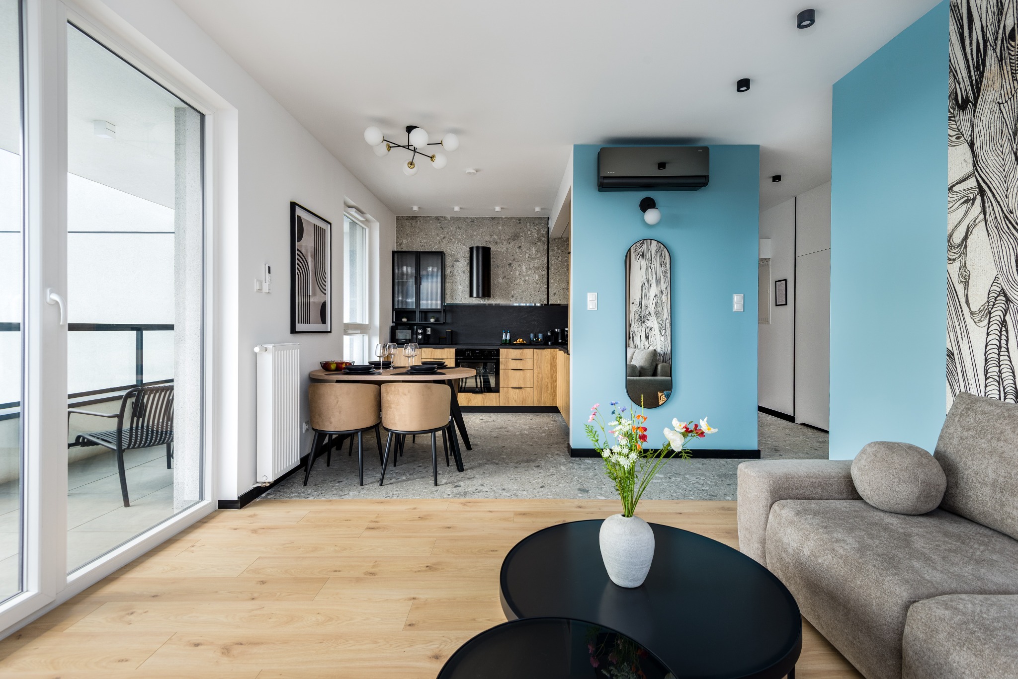 Jasne, przestronne wnętrze mieszkania z otwartą kuchnią, stołem jadalnym i częścią wypoczynkową. Dominuje błękitna ściana i drewniane akcenty. Widok na balkon.
