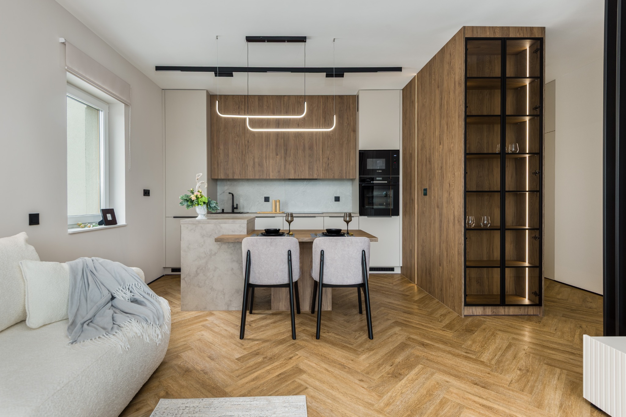 Nowoczesna kuchnia z wyspą i drewnianą zabudową, oświetlona lampami LED. Parkiet w jodełkę, szara sofa z kocem w tle. Minimalistyczny design.