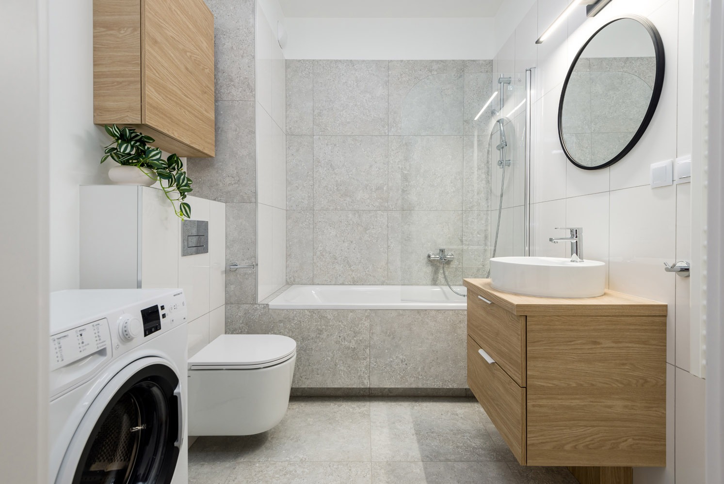 Jasna łazienka z wanną, umywalką na szafce z drewna, toaletą i pralką. Na szafce roślina doniczkowa. Minimalistyczny design, szare płytki i białe ściany.