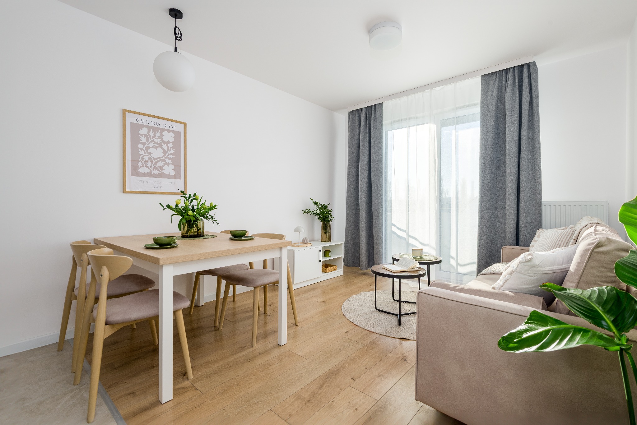 Jasne wnętrze salonu z drewnianą podłogą, stołem jadalnym z kwiatami, beżową sofą i okrągłym dywanem. Minimalistyczny design, naturalne światło.