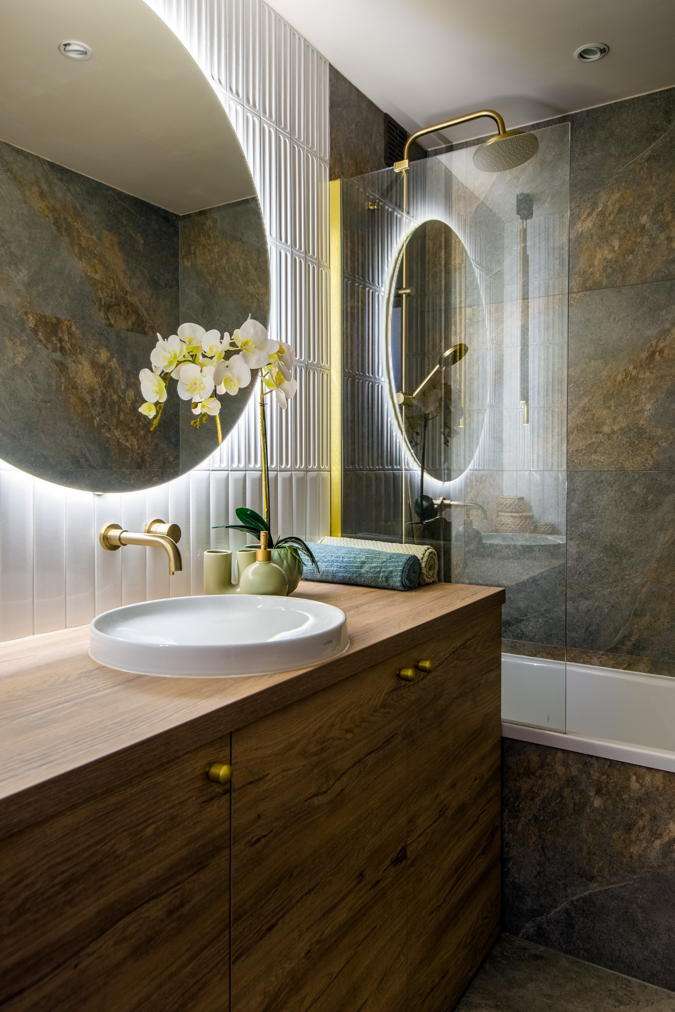 Nowoczesna łazienka z okrągłym lustrem podświetlanym LED, drewnianą szafką pod umywalką i złotą armaturą. Na blacie biała orchidea i zielone akcesoria.