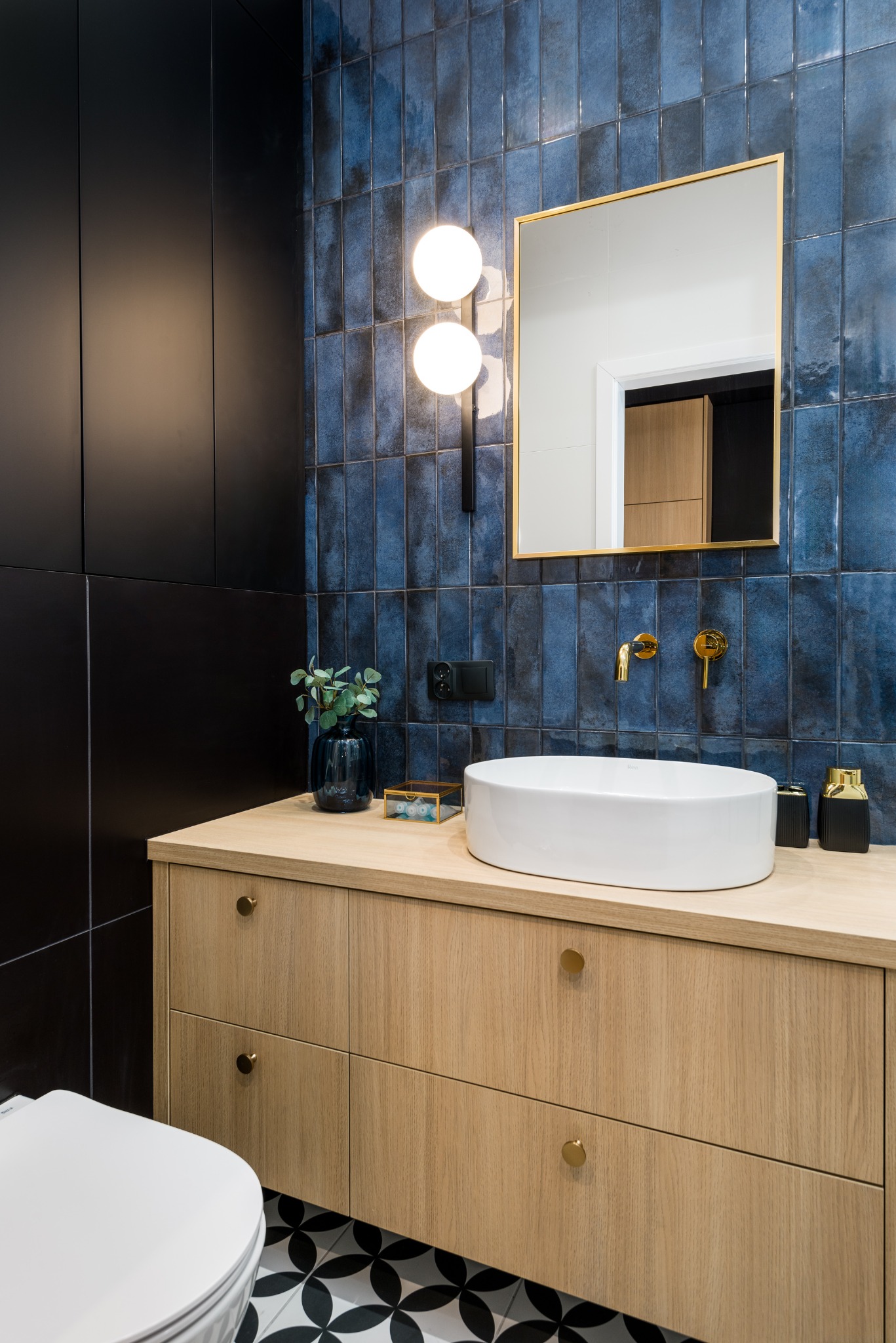 Nowoczesna łazienka z niebieskimi płytkami, drewnianą szafką pod umywalką i złotymi akcentami, oświetlona kulistymi lampami. Minimalistyczny design.
