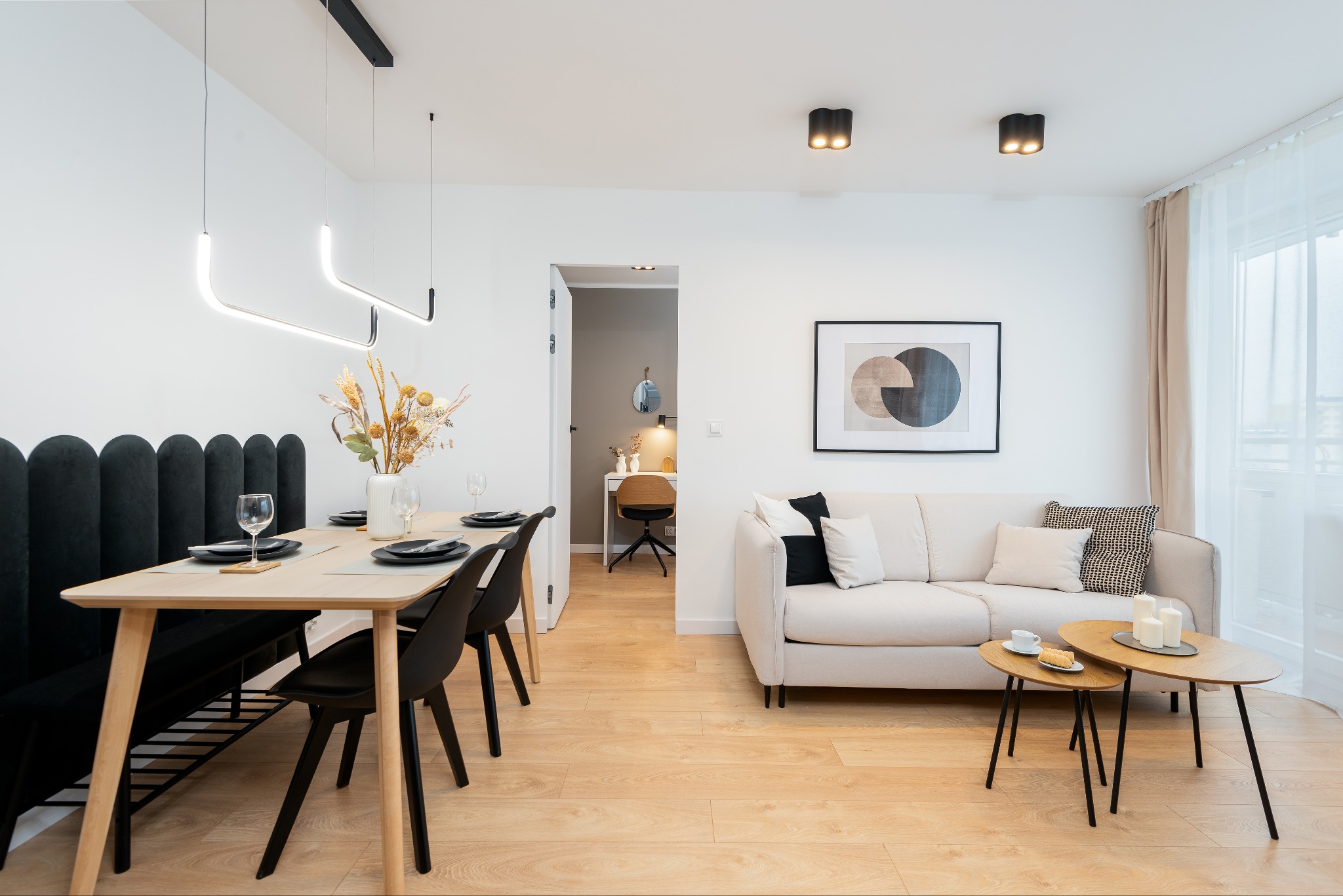 Jasne wnętrze: stół z czarnymi krzesłami i zastawą, sofa z poduszkami, stoliki kawowe z przekąskami, minimalistyczny wystrój, widok na biurko w tle.
