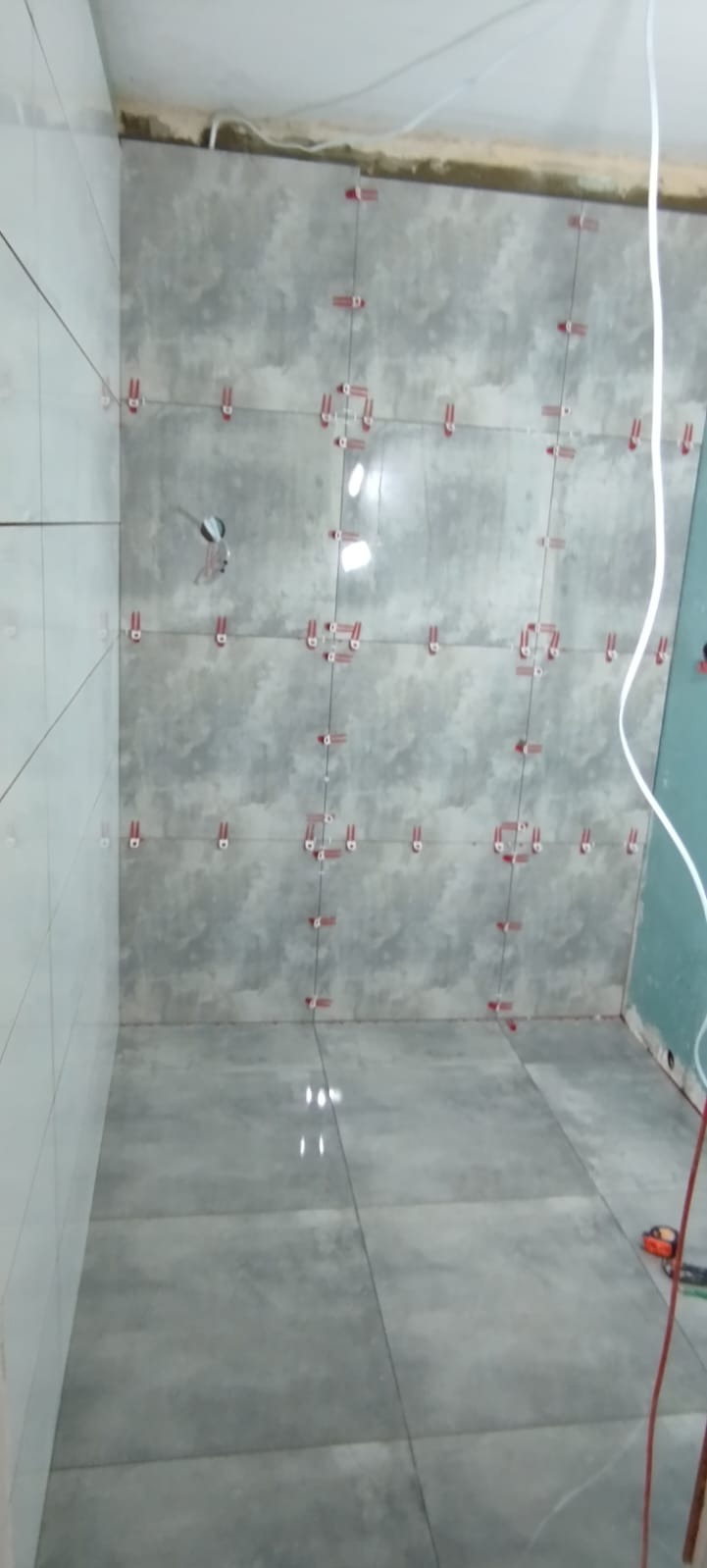 Świeżo ułożone szare płytki na ścianie i podłodze z użyciem czerwonych krzyżyków poziomujących, widoczne kable wystające ze ściany.