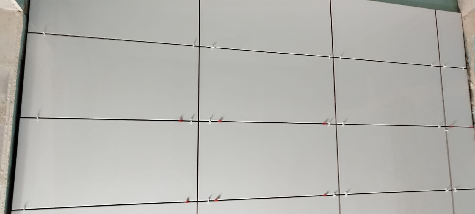 Świeżo ułożone, szare płytki ścienne z użyciem białych i czerwonych krzyżyków dystansowych, widoczne czarne fugi.