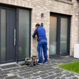 Instalator w niebieskim kombinezonie montuje czujnik alarmowy na elewacji z cegły klinkierowej obok nowoczesnych okien i drzwi, z torbą narzędziową na ziemi.