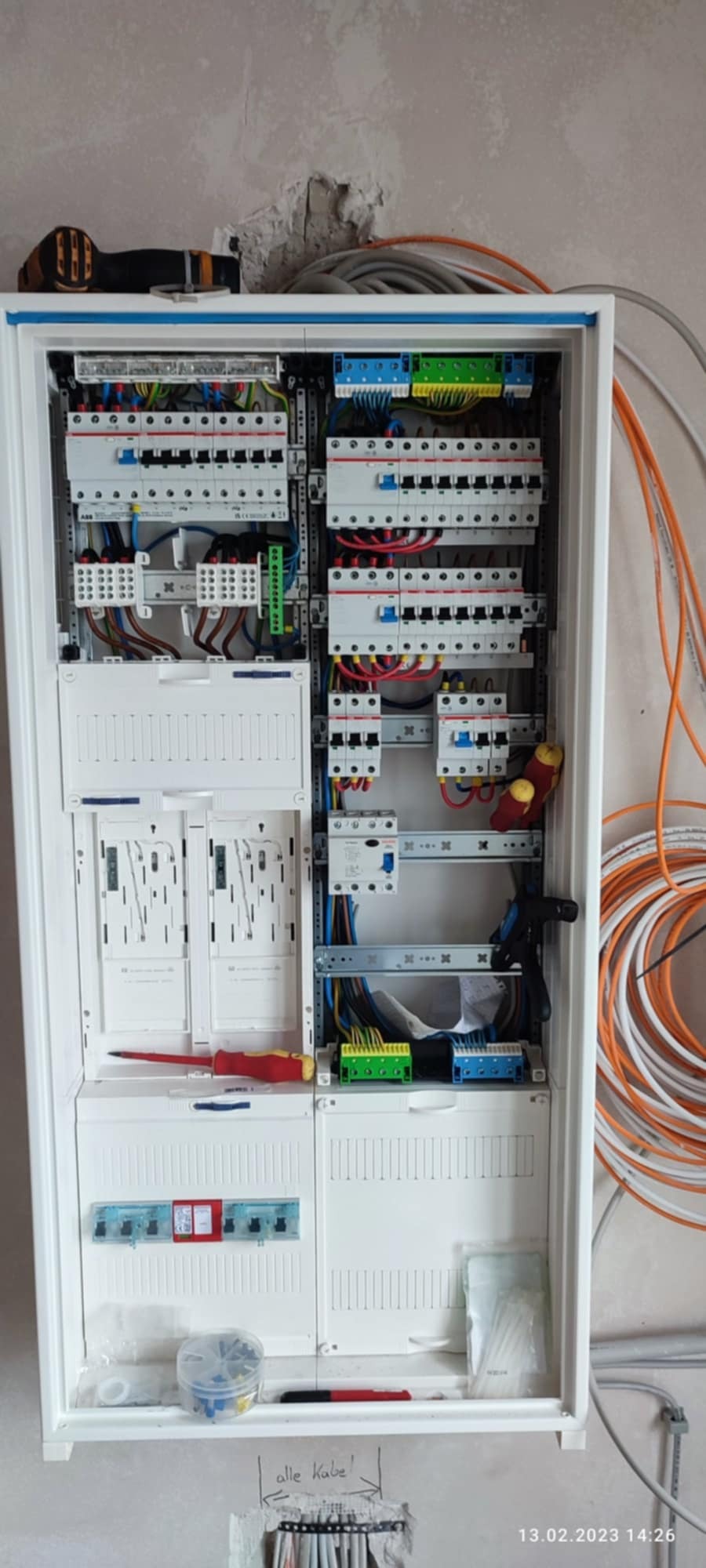 Otwarta skrzynka rozdzielcza z widocznymi bezpiecznikami, przewodami i elementami montażowymi; w tle surowa ściana i zwój pomarańczowych kabli.