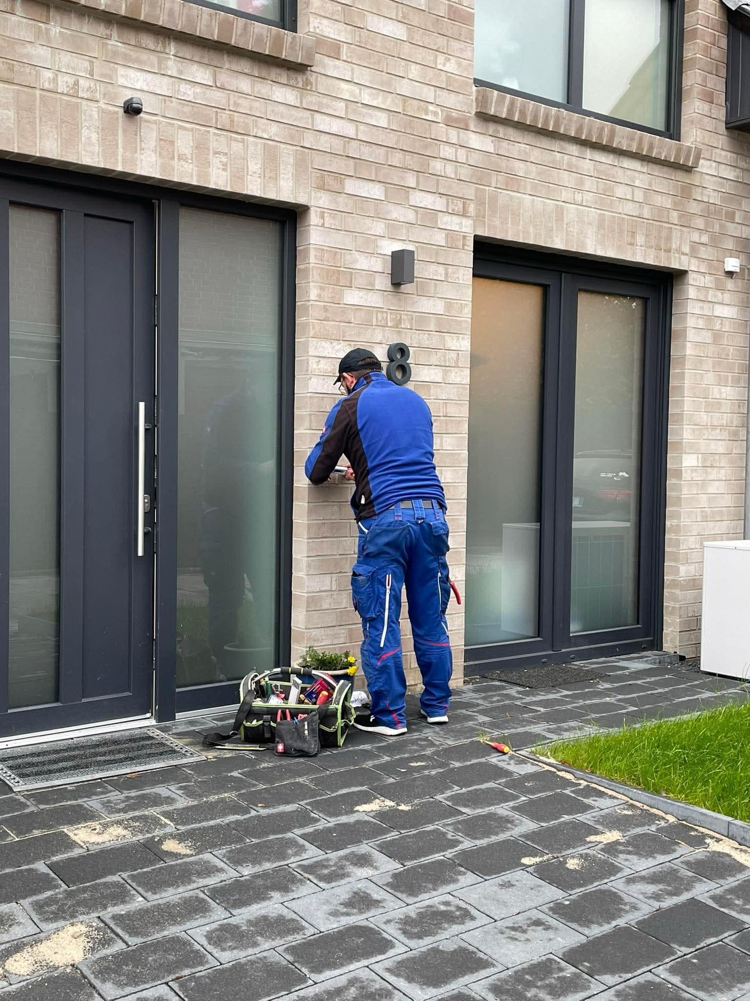 Instalator w niebieskim kombinezonie montuje czujnik alarmowy na elewacji z cegły klinkierowej obok nowoczesnych okien i drzwi, z torbą narzędziową na ziemi.