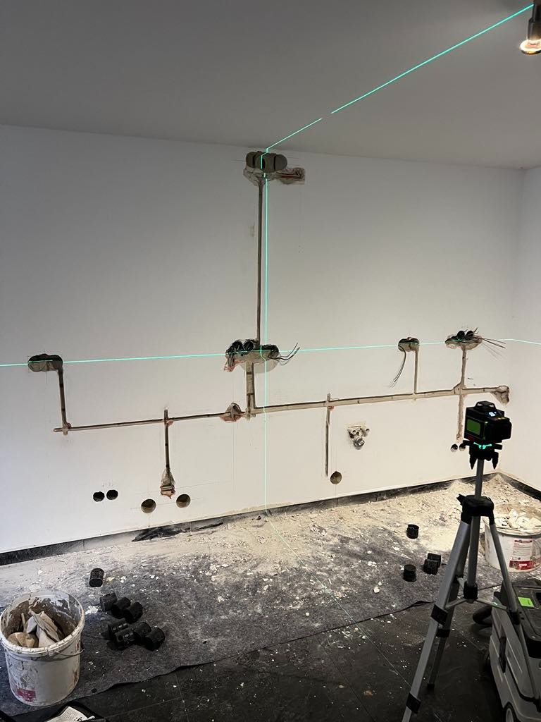 Wnętrze w trakcie prac elektrycznych: widoczne przewody w ścianie, puszki instalacyjne, poziomica laserowa na statywie, oraz bałagan na podłodze w postaci gruzu i resztek materiałów budowlanych.